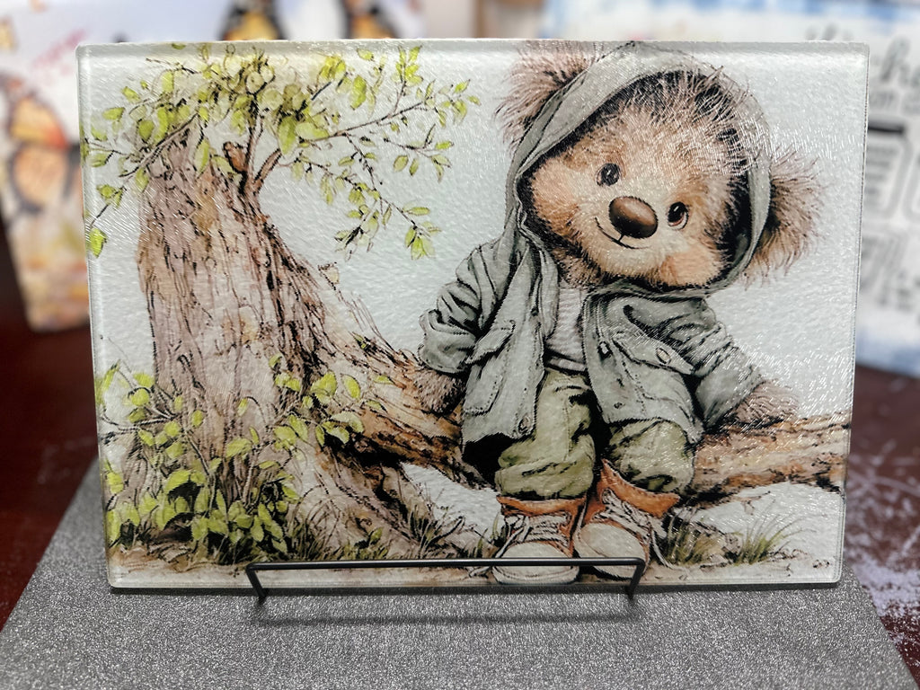 Decorative plaque with a teddy bear in a coat and hat sitting on a log, surrounded by nature.